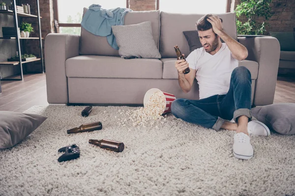 Foto do cara de raça mista sentado tapete inclinado sofá segurar garrafa de cerveja pipoca no chão tinha entretenimento louco após a festa sofrem ressaca manhã dor de cabeça confuso apartamento dentro de casa — Fotografia de Stock