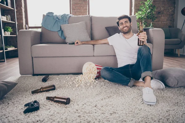 Foto de tipo de raza mixta sentado alfombra cerca del sofá sosteniendo botella de cerveza palomitas de maíz piso divertirse despedida de soltero loco no importa sucio piso sucio reunión mejores amigos invitados en el interior — Foto de Stock