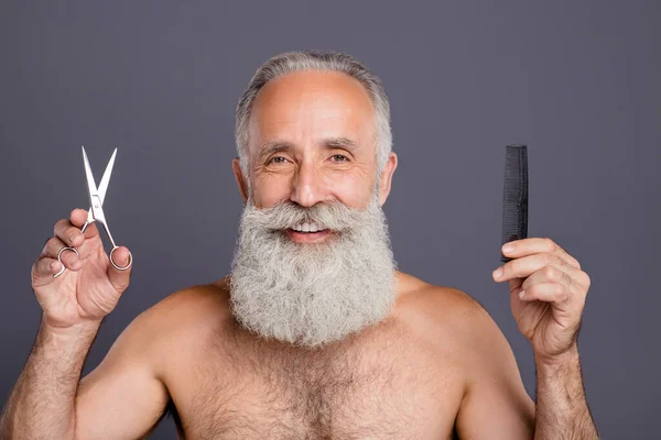 Фото веселого позитивного зрелого мужчины с ножницами и расчёской для волос, улыбающегося зубами, осознающего важность ухода за волосами изолированного серого цвета фона — стоковое фото