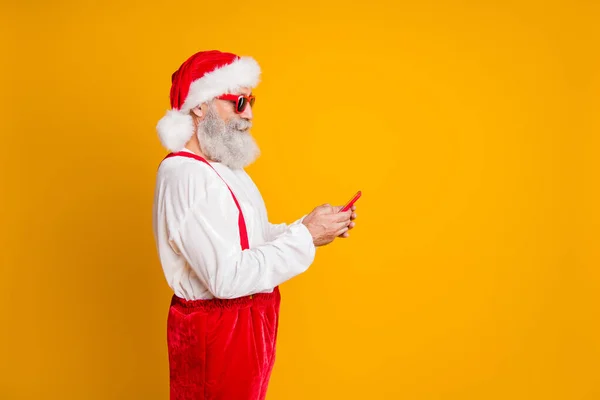 Profil côté photo de graves cheveux gris grand-père hipster en rouge santa claus chapeau utiliser smartphone recherche x-mas saison nouvelle année veille temps réductions type sms isolé couleur jaune fond — Photo