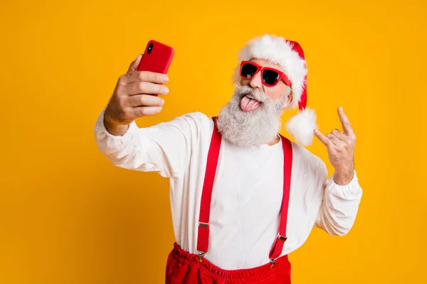 Χ-μας πάρτι σκληρά. Πορτρέτο του αστείου τρελό santa claus σε κόκκινο καπέλο λάβει selfie έξυπνο τηλέφωνο Χριστούγεννα blogger δείχνουν κέρατα γκριμάτσα γλώσσα έξω φορούν τιράντες πουκάμισο απομονωμένο κίτρινο χρώμα φόντο — Φωτογραφία Αρχείου