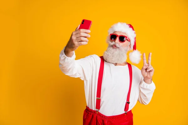 Portrait of funky grey white hair santa claus in red hat cap enjoy x-mas christmas time take selfie smartphone make v-sign send air kiss wear shirt suspenders isolated yellow color background — Stock Photo, Image