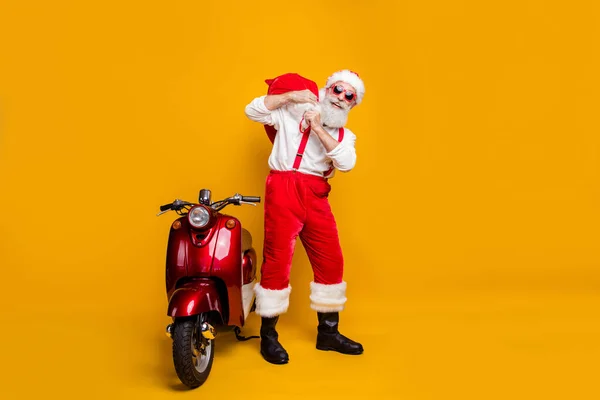 Foto de longitud completa de Santa Claus loco divertido en sombrero rojo llevar gran saco x-mas tradición Navidad partido stand motocicleta desgaste tirantes aislado color amarillo fondo —  Fotos de Stock