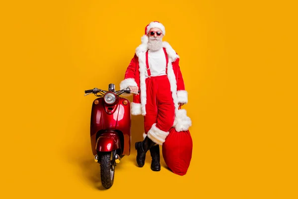 Full size photo of confident grandfather in red santa claus hat true driver rider hold big sack x-mas dream gift stand near scooter wear shirt suspenders isolated yellow color background — Stock Photo, Image