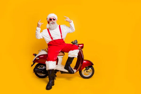 Foto de cuerpo completo de Santa Claus pelo blanco loco en el estado de ánimo festivo equitación fiesta de x-mas sentado en bicicleta mostrando cuernos usar gafas de sol pantalones gorra botas aisladas de color amarillo fondo —  Fotos de Stock
