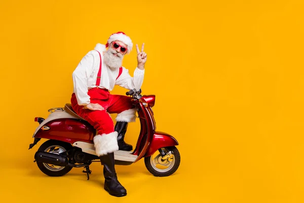 Foto de cuerpo completo de Santa Claus blanco funky listo para x-mas fiesta temática sentado bicicleta mostrando v-signo de desgaste hipster sol especificaciones pantalones gorra botas aislado color amarillo fondo —  Fotos de Stock