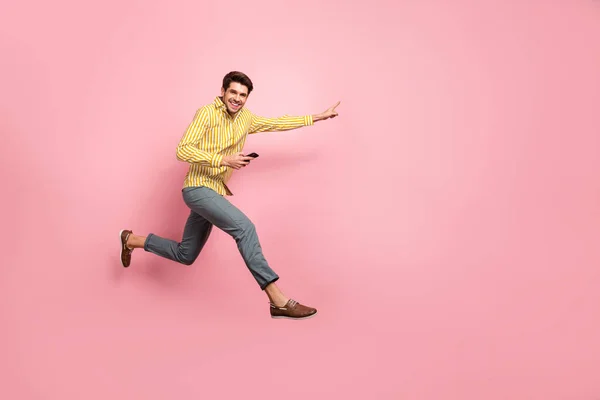 Full length profile photo of beautiful guy jump high hold phone rush shopping indicate finger κενό χώρο πώληση φορούν ριγέ πουκάμισο παντελόνι απομονωμένο ροζ χρώμα φόντο — Φωτογραφία Αρχείου