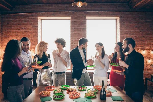 Foto de pessoas alegres vestindo formalmente de bom humor em pé à mesa colocada com comida festiva segurando copos de bebida alcoólica falando — Fotografia de Stock