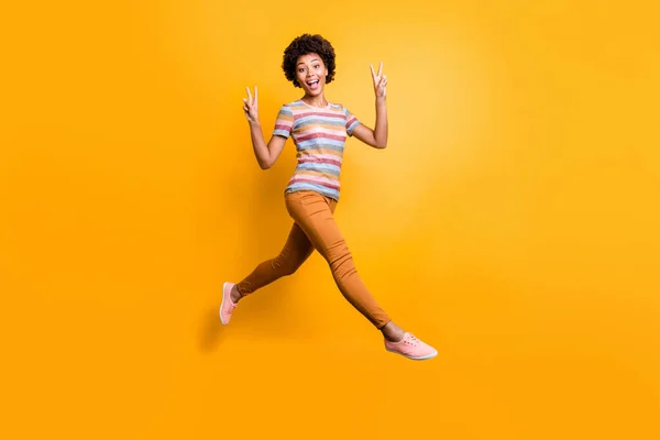 Полная длина фото позитивной веселой темнокожей девушки весело провести весенние праздники чувствую себя фанки смешно сумасшедший прыжок сделать V-знак носить полосатую футболку коричневые брюки брюки изолированный желтый цвет фона — стоковое фото