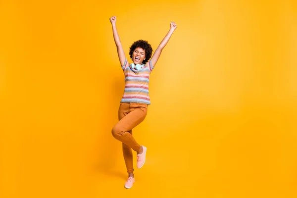 Full size foto di estatica pazzo afro ragazza americana hanno auricolare trovato canzone preferita radio ascoltare musica alzare i pugni urlare indossare a strisce t-shirt marrone pantaloni pantaloni isolato colore brillante sfondo — Foto Stock