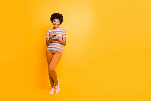 Full length photo of positive chappy afro american girl stand use smartphone type sms Η φίλη της στο δίκτυο μέσων κοινωνικής δικτύωσης φοράει ριγέ t-shirt καφέ παντελόνι απομονωμένο κίτρινο χρώμα φόντο — Φωτογραφία Αρχείου