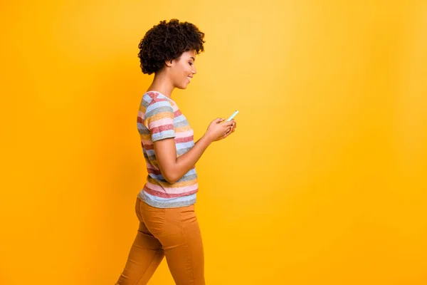 Profile side photo of positive cheerful afro american girl use cell type blog post comment enjoy read feed news go walk wear striped t-shirt brown pants pants isolated yellow color background — Zdjęcie stockowe