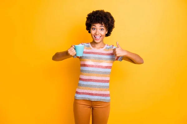 Utmärkt kvalitet arabica. Porträtt av glada förvånad afro amerikansk flicka hålla mugg dryck visa tumme upp annonsera promo bära randig t-shirt bruna byxor isolerad gul färg bakgrund — Stockfoto