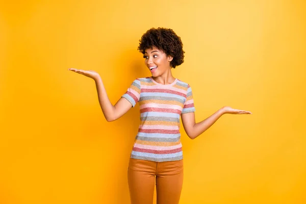 Photo of amazing dark skin curly model lady holding two new products on palms advising cheap one wear casual striped t-shirt pants isolated on yellow background — Stock Photo, Image