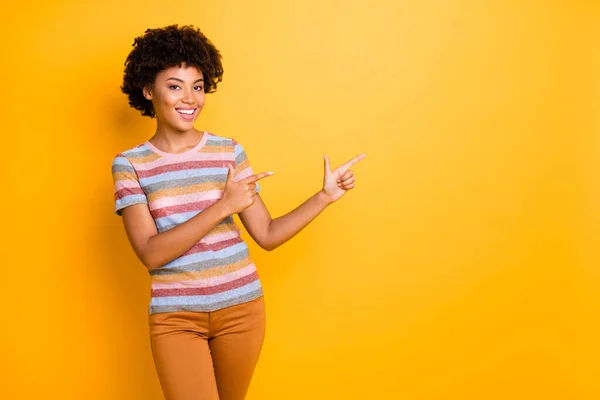 Foto de la increíble señora de piel oscura que indica los dedos espacio vacío que aconseja nuevo producto fresco desgaste casual camiseta de rayas y pantalones aislados en el fondo amarillo —  Fotos de Stock