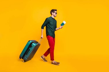 Full length body size side profile photo of man carrying his baggage holding passport and tickets to get on board walking towards airplane in red pants trousers isolated vivid color yellow background clipart