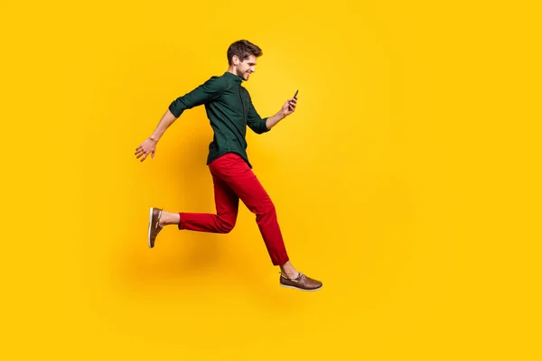 Volledige lengte profiel zijkant foto van positieve vrolijke man gebruik smartphone zoeken kortingen op social media springen run snel dragen groen shirt rood broek geïsoleerde gele kleur achtergrond — Stockfoto