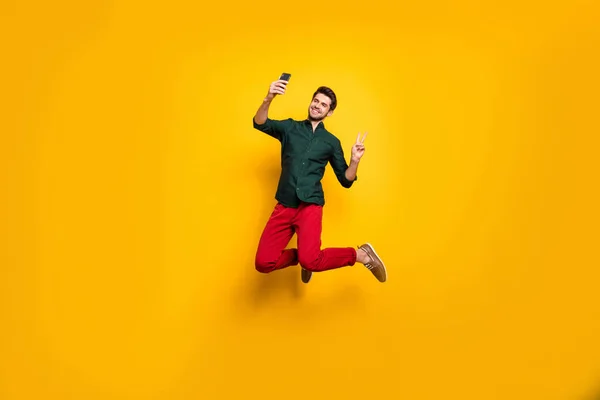 Plná délka tělo velikost fotografie veselý pozitivní chlap ukazuje V-znamení přičemž selfie na sobě zelené tričko červené kalhoty kalhoty s úsměvem zubatě skákání nahoru izolované živé barevné pozadí — Stock fotografie