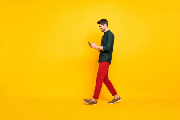 Volledige lichaamslengte foto van bewegende vrolijke wandelen verslaafde aantrekkelijke man bladeren door zijn telefoon dragen rode broek schoenen geïsoleerd over geel levendige kleur achtergrond — Stockfoto