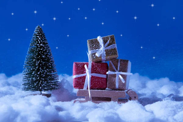 Foto av lilla leksak Santa claus slädar bär gula röda bruna presentaskar för jul jul fest stå nära evergreen tall under natten blå himmel bakgrund — Stockfoto