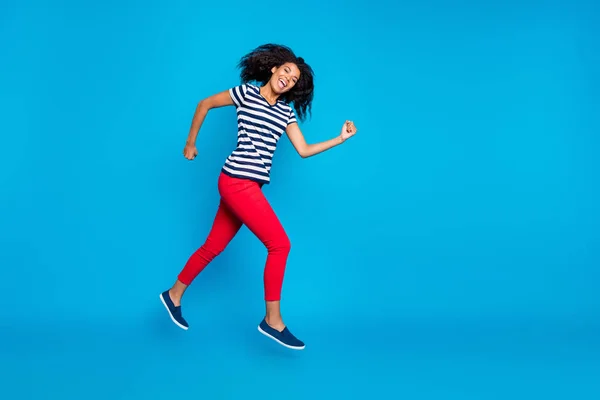 Perfil de comprimento total foto lateral de alegre louco afro americano menina salto correr rápido após descontos de primavera usar listrado t-shirt calças vermelhas calças tênis isolado sobre fundo de cor azul — Fotografia de Stock