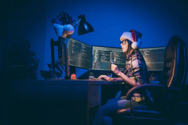 Профиль фото программистки с игристым бокалом вина, смотрящей на экран, работающий допоздна, встречает новогоднюю встречу в одиночестве в темном офисе трудоголика носить шапочку Санта в помещении — стоковое фото