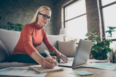 Photo of blond business lady texting notebook colleagues working home noticing startup details in organizer wear specs orange pullover sitting comfy sofa indoors clipart
