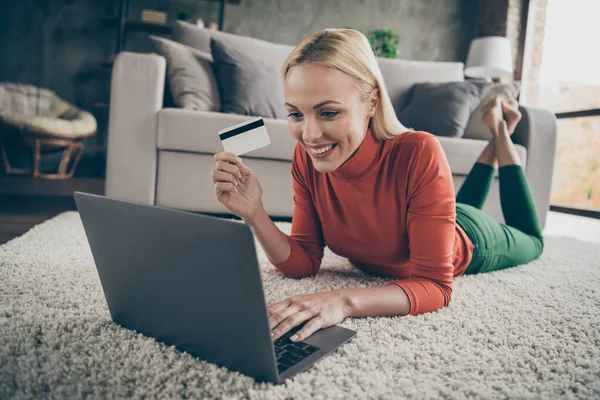 照片上漂亮的家庭主妇在网上选择笔记本电脑购买准备好的信用卡付款地毯地板靠近沙发休闲装客厅在室内 — 图库照片