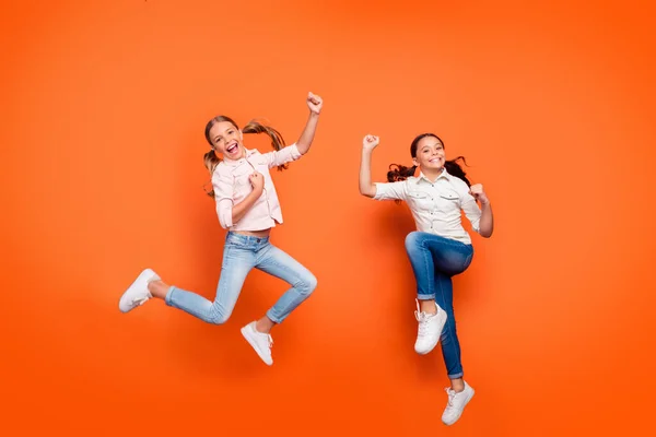 Pleine longueur photo de positif réjouissant deux copines saut repos détente gagner école sport compétition lever poings porter chemise blanche jeans en denim isolé sur fond de couleur orange — Photo