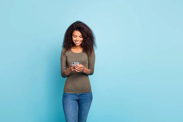 Porträtt av positiva glada fokuserad bloggare mulatto flicka med hjälp av sin smartphone läsa feednews typ feedback på bloggar bära grön tröja denim jeans isolerad över blå färg bakgrund — Stockfoto