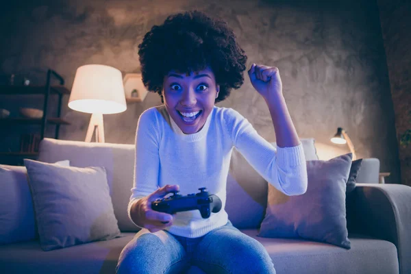 Ritratto di deliziato divertente afro americano ragazza tenere joystick giocare video gioco gara vincere alzare i pugni urlare sì sedersi accogliente divano in casa stanza serale al chiuso — Foto Stock