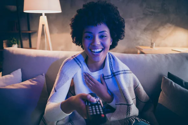 Portrait of positive funny afro american girl enjoy evening time watch comedy laugh hold remote control switch tv channel sit couch under lamp cover checkered soft blanket in house indoors — Stock Photo, Image