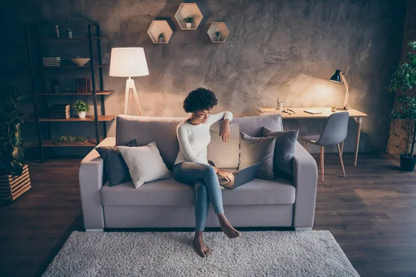 Foto de vista de ángulo alto de piel oscura increíble señora rizada navegación portátil mensajes de texto con amigos sentado sofá acogedor desgaste casual suéter jeans ropa noche plana en el interior — Foto de Stock