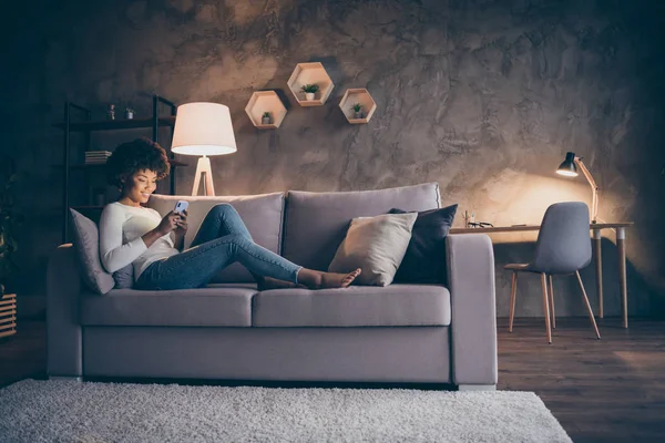 Profile photo of pretty dark skin curly lady domestic atmosphere texting telephone lying comfy couch in lamp light wear casual outfit evening living room indoors