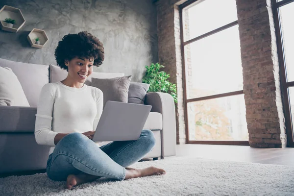 Foto di incredibile pelle scura riccia signora navigazione utilizzando notebook sms con gli amici seduti accogliente pavimento vicino divano indossare casual maglione jeans vestito piatto al chiuso — Foto Stock