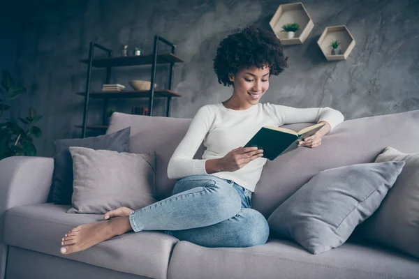 Foto de atractiva mujer rizada de piel oscura sosteniendo las manos favoritas de la novela de amor histórico leyendo emocionado mejor momento sentado acogedor sofá desgaste casual suéter jeans traje plano en el interior — Foto de Stock