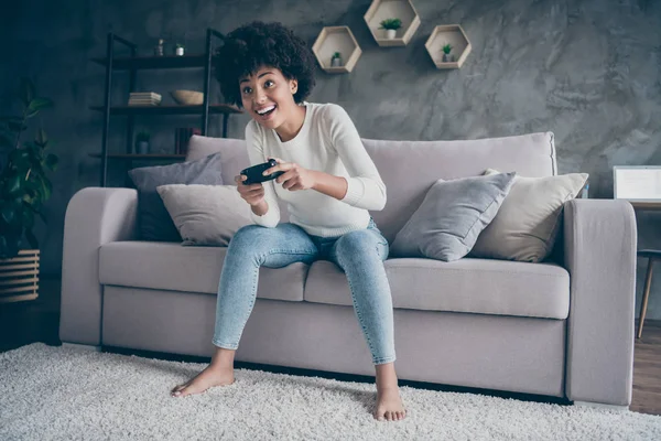 Fénykép a vicces sötét bőr göndör hölgy játszik videojátékok addicted akar nyerni hold joystick ül kényelmes kanapé alkalmi pulcsi farmer ruha lakás beltéri — Stock Fotó