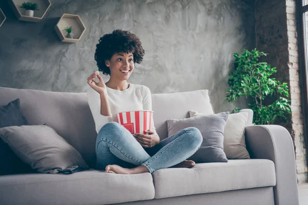 Foto di abbastanza divertente pelle scura ondulata signora umore familiare mangiare popcorn guardare preferito umorismo show televisivo seduto accogliente divano casual maglione jeans vestito piatto al coperto — Foto Stock