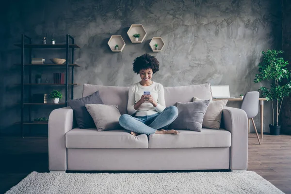 Foto de bonita piel oscura rizado señora hogareño ambiente doméstico mensajes de texto teléfono con amigos lectura instagram puestos sentado cómodo sofá traje casual loft plano interior — Foto de Stock