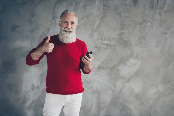 Mutlu, beyaz sakallı, cep telefonu kullanan yaşlı bir adam kış sezonu indirimlerini onaylıyor. Noel 'de Noel reklamlarında baş parmağında kırmızı kazak, beyaz pantolon, gri arka plan. — Stok fotoğraf