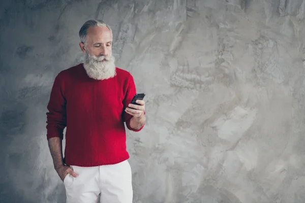 男性の深刻な引退した老人の肖像画スタンドコピースペース使用スマートフォン読み取りX-masクリスマス割引着用赤トレンディーなプルオーバーホワイトパンツ孤立グレー具体的な壁の色の背景 — ストック写真