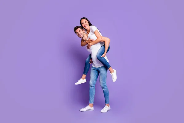 Gedraaide volledige lichaamslengte foto van schattige paar van twee geliefde mensen meeliften in wit t-shirt glimlachen getand met man dragen zijn vrouw geïsoleerde violet pastel kleur achtergrond — Stockfoto