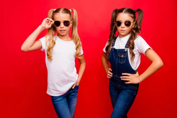 Foto av två ganska små damer långa svansar självförtroende tittar på kameran bära cool sol specs jeans övergripande vit t-shirt isolerad röd ljus färg bakgrund — Stockfoto