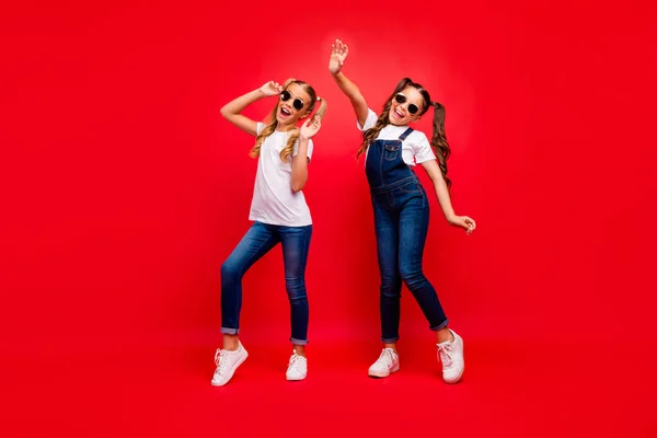 Full längd foto av två ganska små damer långa svansar glädje på skolfesten bästa vänner bära cool sol specs jeans övergripande vit t-shirt isolerad röd ljus färg bakgrund — Stockfoto
