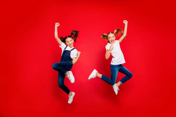 Volledige foto van vrolijke twee kleine mensen vriendinnen vieren triomf hef vuisten schreeuwen ja spring dragen witte t-shirt denim jeans sneakers geïsoleerde heldere kleur achtergrond — Stockfoto