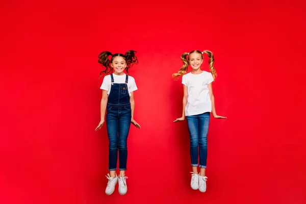 Foto de corpo inteiro de duas pequenas namoradas positivas saltar desfrutar de fins de semana de primavera tempo de lazer desgaste branco t-shirt jeans macacão tênis isolado cor vermelha fundo — Fotografia de Stock