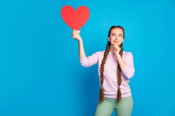 Portrait d'une jeune fille intéressée réfléchie avec de longues queues de cochon tenir carte de papier rouge cadeau de coeur pour la Saint-Valentin penser à la date Cupidon porter pull rose pantalon vert isolé fond de couleur bleue — Photo