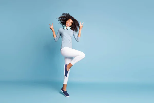 Longitud completa tamaño del cuerpo foto de alegre positivo bonito joven saltando bailando que muestra v-signo en el calzado pantalones blancos aislados pastel fondo de color azul —  Fotos de Stock