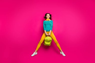 Full length body size photo of pretty sweet charming curly cute girlfriend wearing yellow pants trousers white footwear smiling toothily jumping up with rucksack isolated vivid fuchsia background clipart