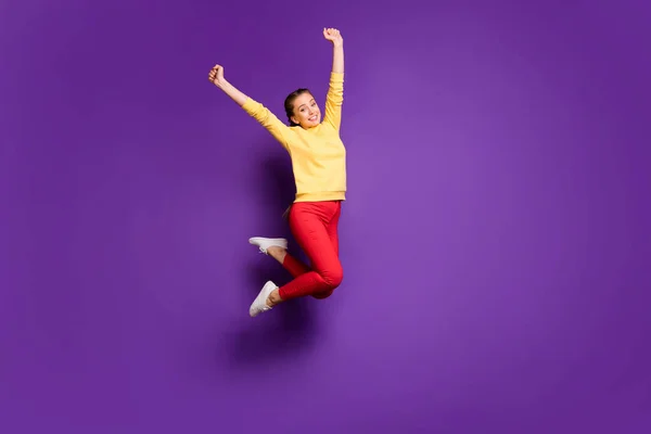 Teljes hosszúságú fotó csinos tini hölgy jumping magas ünneplő nyaralás idő felemeli a kezét viselni alkalmi sárga pulóver piros nadrág elszigetelt lila szín háttér — Stock Fotó